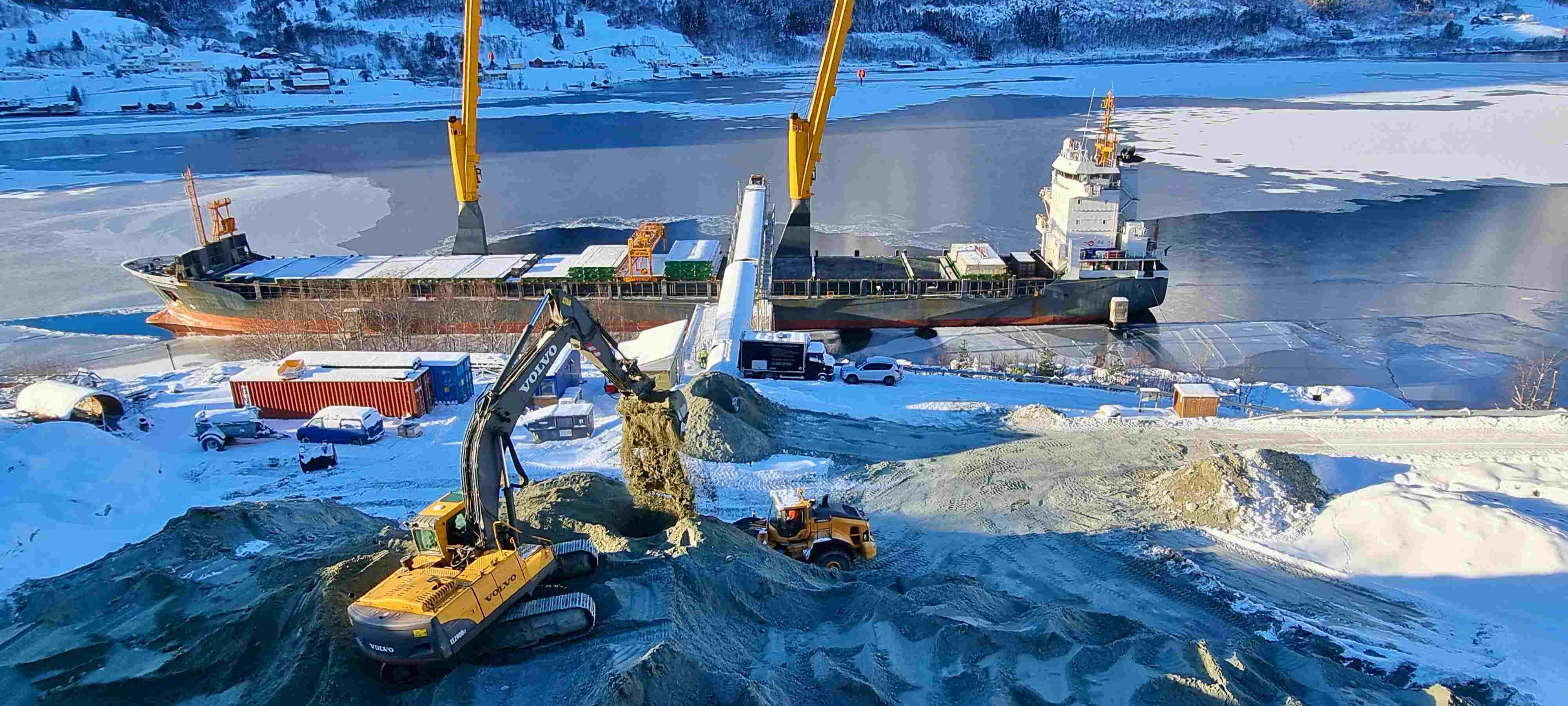En vinterlig havn med en stor fraktbåt til kai. Maskiner og lastebiler er synlige på land, der sand eller grus blir lastet fra et stort haug. Is dekkes av deler av vannet.