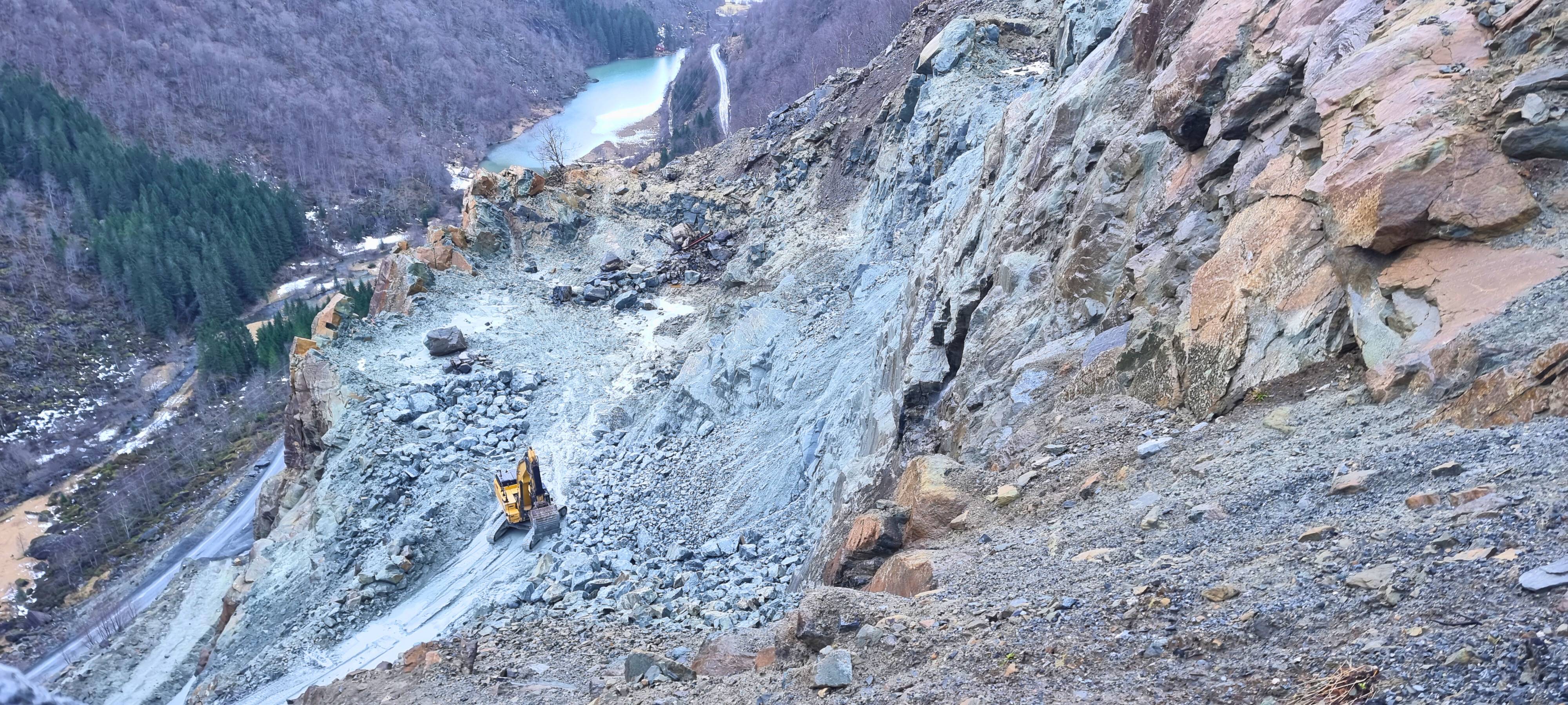  En gravemaskin arbeider på et skrått område. Landskapet viser brune og grå steiner med et frustrerende utsikt over en dal og en elv i bakgrunnen.