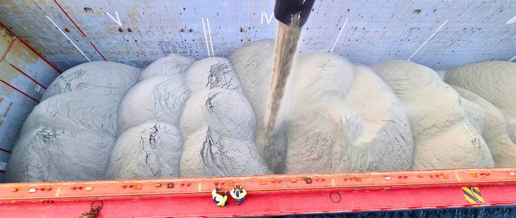 En oversikt over en fraktbåt som laster sand. To personer står på bunnen av båten, mens en rørledning leverer sand direkte inn i lasten. Båten er delvis fylt med sand i bølger.